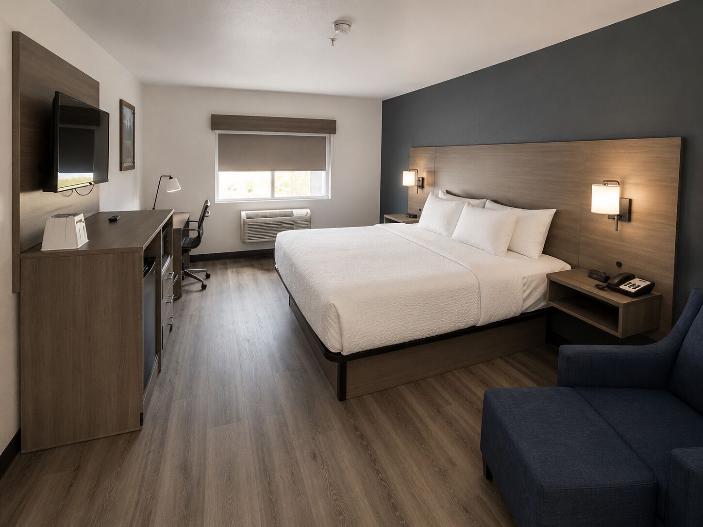 A hotel room with a bed, bedside table, chair, desk, TV, and a window with blinds.