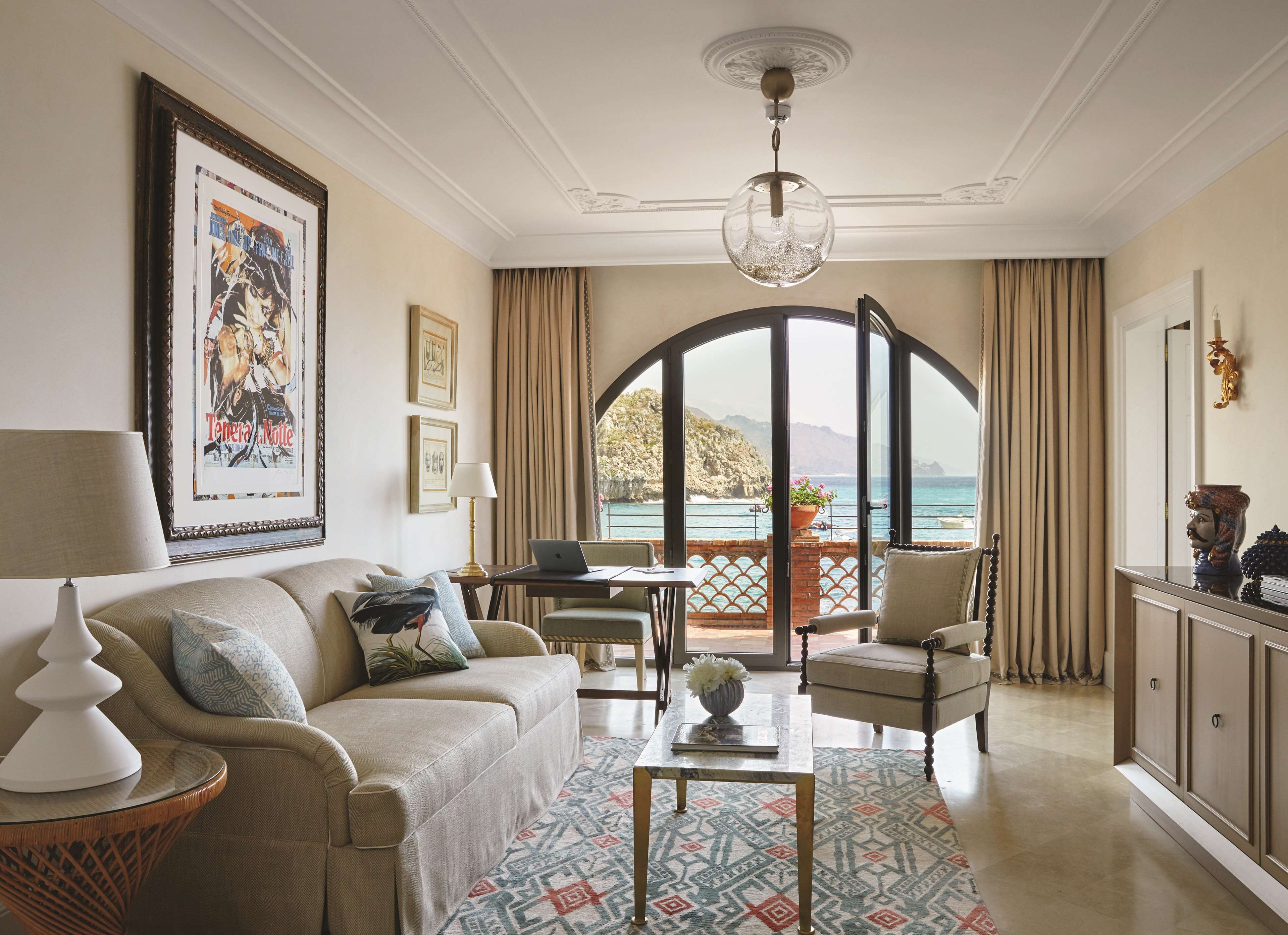 A living room with a sofa, armchairs, a coffee table, a laptop, and a view of the sea through a large arched window.