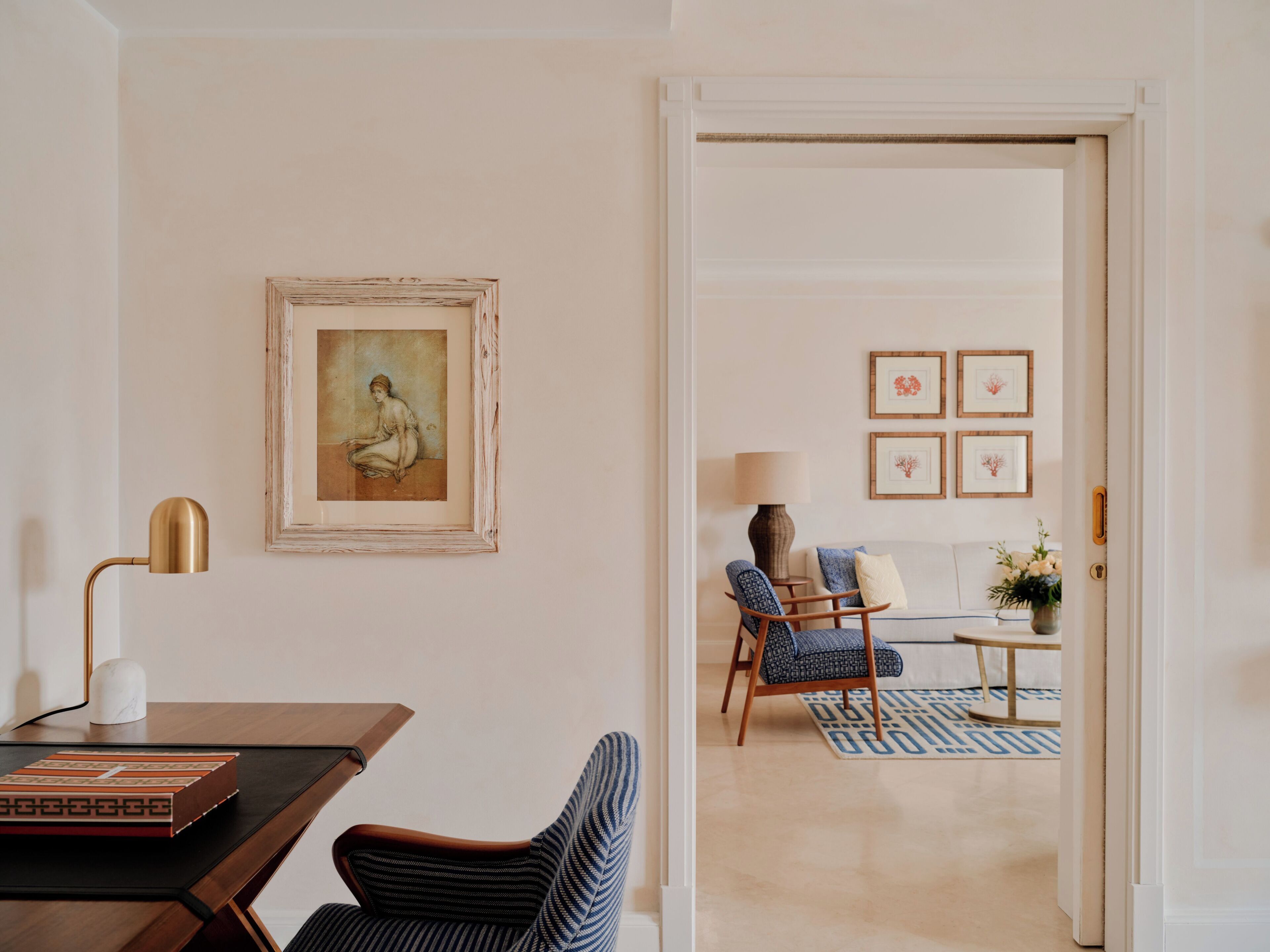 Una habitación con escritorio, silla, lámpara, una obra de arte enmarcada y vistas a otra estancia con sofá y sillones.