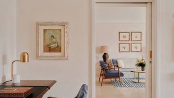 Una habitación con escritorio, silla, lámpara, una obra de arte enmarcada y vistas a otra estancia con sofá y sillones.