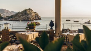 An outdoor dining area with tables and chairs, overlooking a body of water with boats and a rocky island. - Villa Sant'Andrea, A Belmond Hotel, Taormina Mare (Taormina)