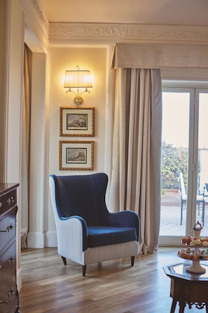 A room with a blue armchair, a wall-mounted lamp, two framed pictures, a wooden dresser, and a view through a glass door.