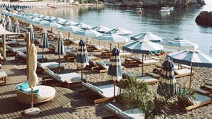 A beach with lounge chairs and umbrellas, a wooden deck, and a view of the sea and a rocky island. - Villa Sant'Andrea, A Belmond Hotel, Taormina Mare (Taormina)