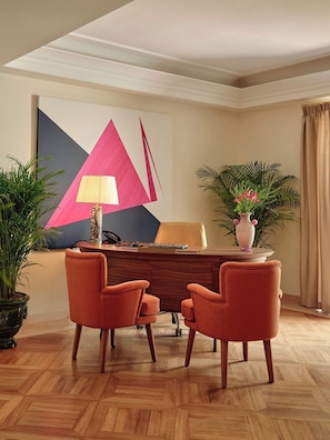 A conference room with a wooden desk, two orange chairs, a lamp, a potted plant, and a large abstract painting on the wall.