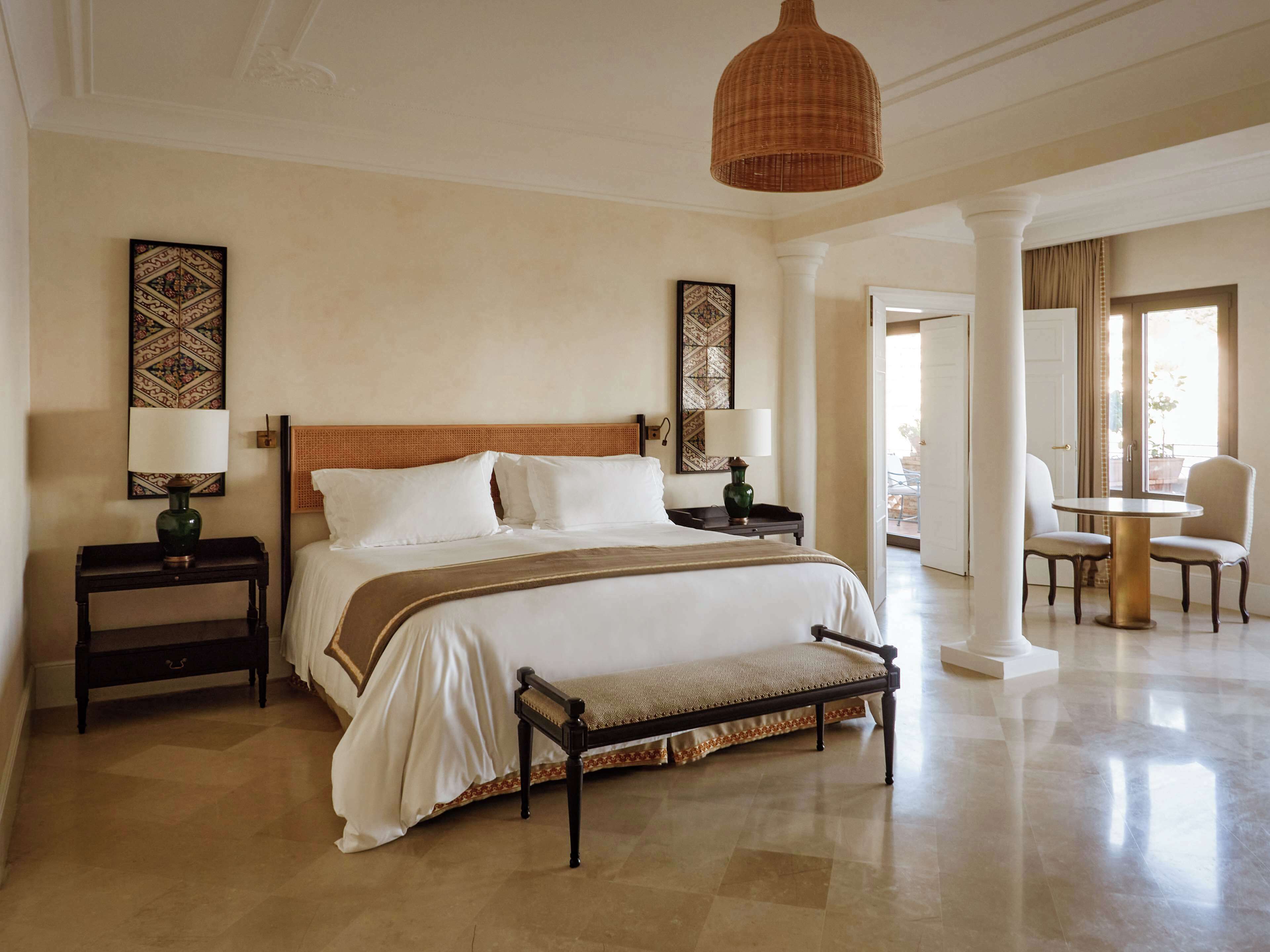 A hotel room with a large bed, two bedside tables, a bench, and a dining area with a table and chairs.