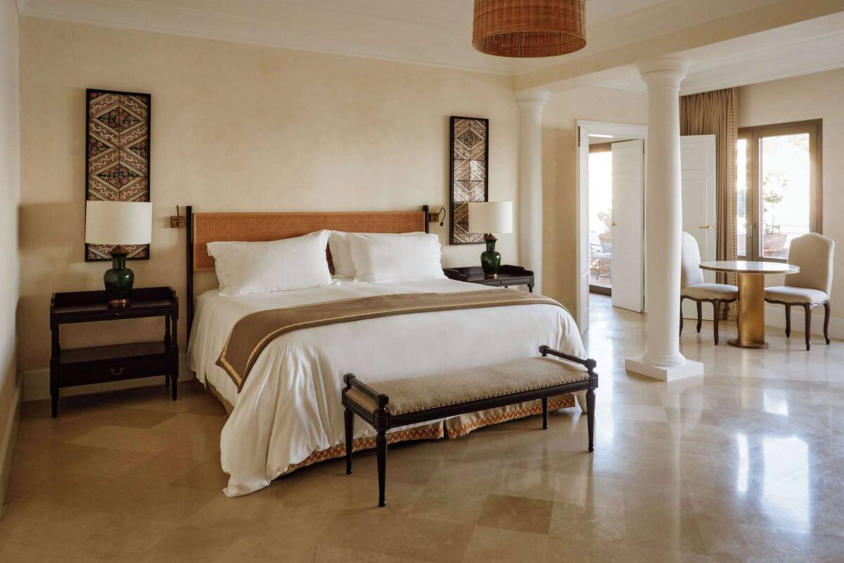Une chambre d’hôtel avec un grand lit, deux tables de chevet, un banc et un coin repas comprenant une table et des chaises.