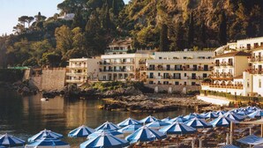 A coastal hotel with multiple balconies overlooking the sea, a stone wall, and numerous blue and white striped umbrellas. - Villa Sant'Andrea, A Belmond Hotel, Taormina Mare (Taormina)