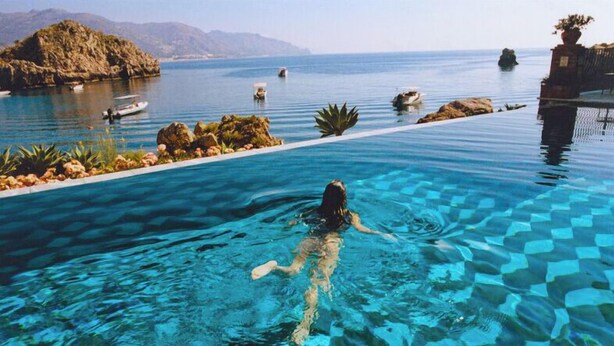 A person swimming in a clear blue infinity pool with a view of the sea and rocky outcrops. - Villa Sant'Andrea, A Belmond Hotel, Taormina Mare (Taormina)