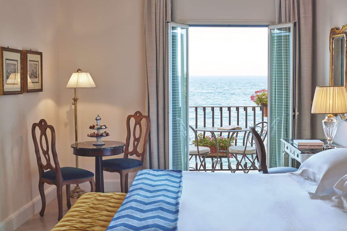 Une chambre d’hôtel avec un lit, une petite table avec des chaises, un balcon donnant sur la mer et une fenêtre avec des rideaux.