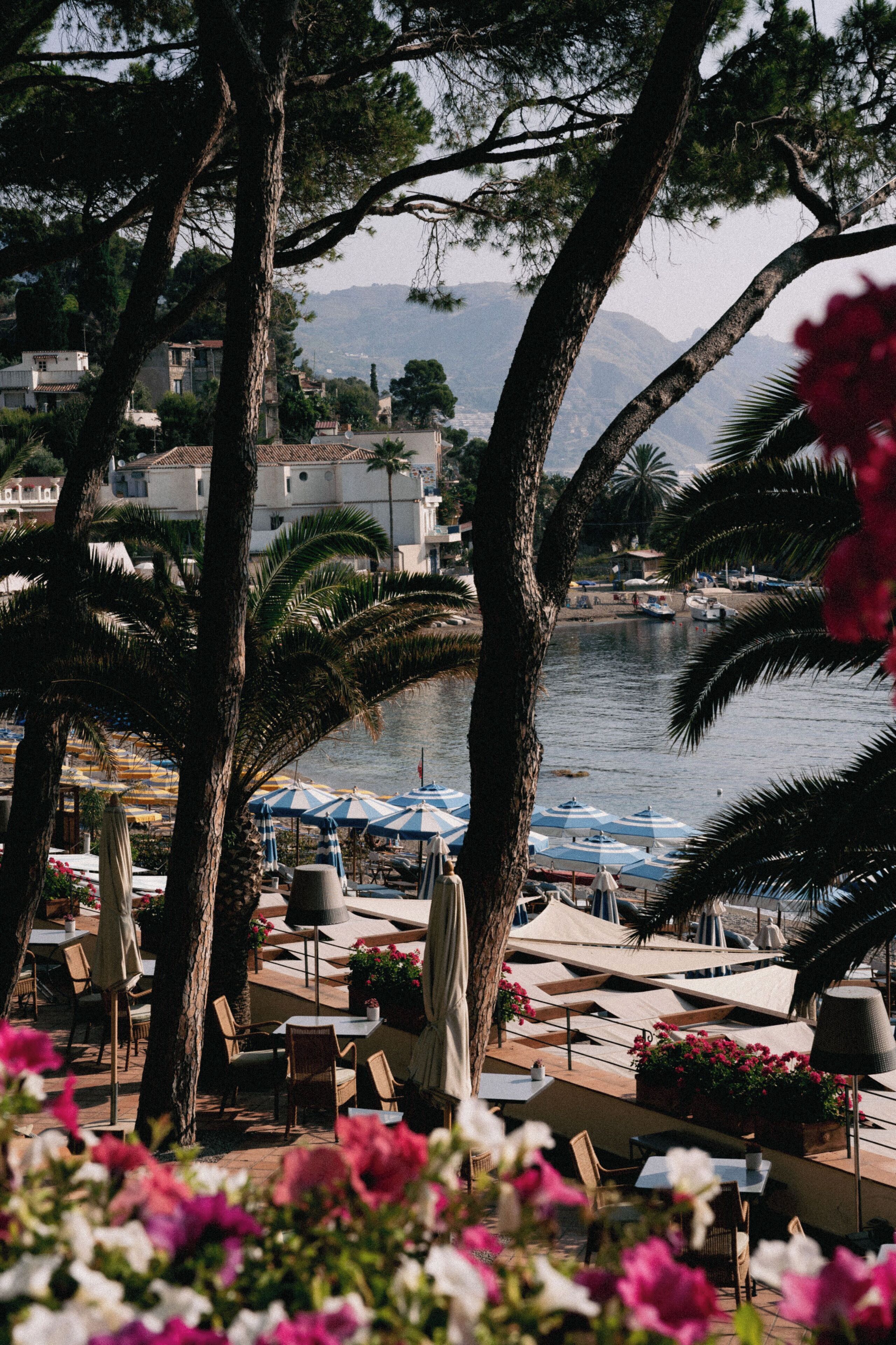 Un espace extérieur aménagé avec des parasols, des tables et des chaises, situé en bord de mer et offrant une vue sur une étendue d’eau et des bâtiments en arrière-plan.