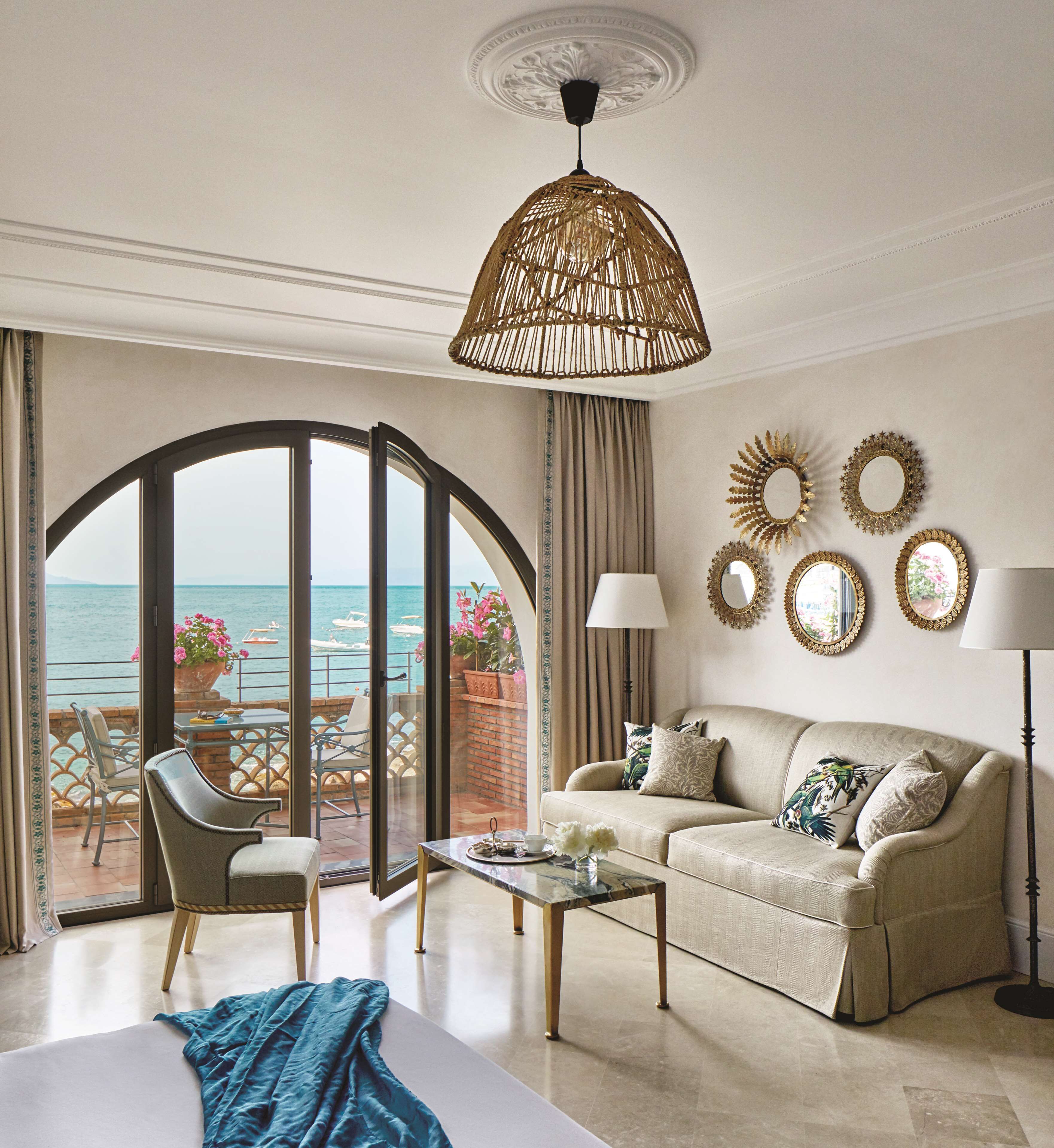A spacious living room with a sofa, armchairs, and a coffee table. A large window offers a view of the sea.