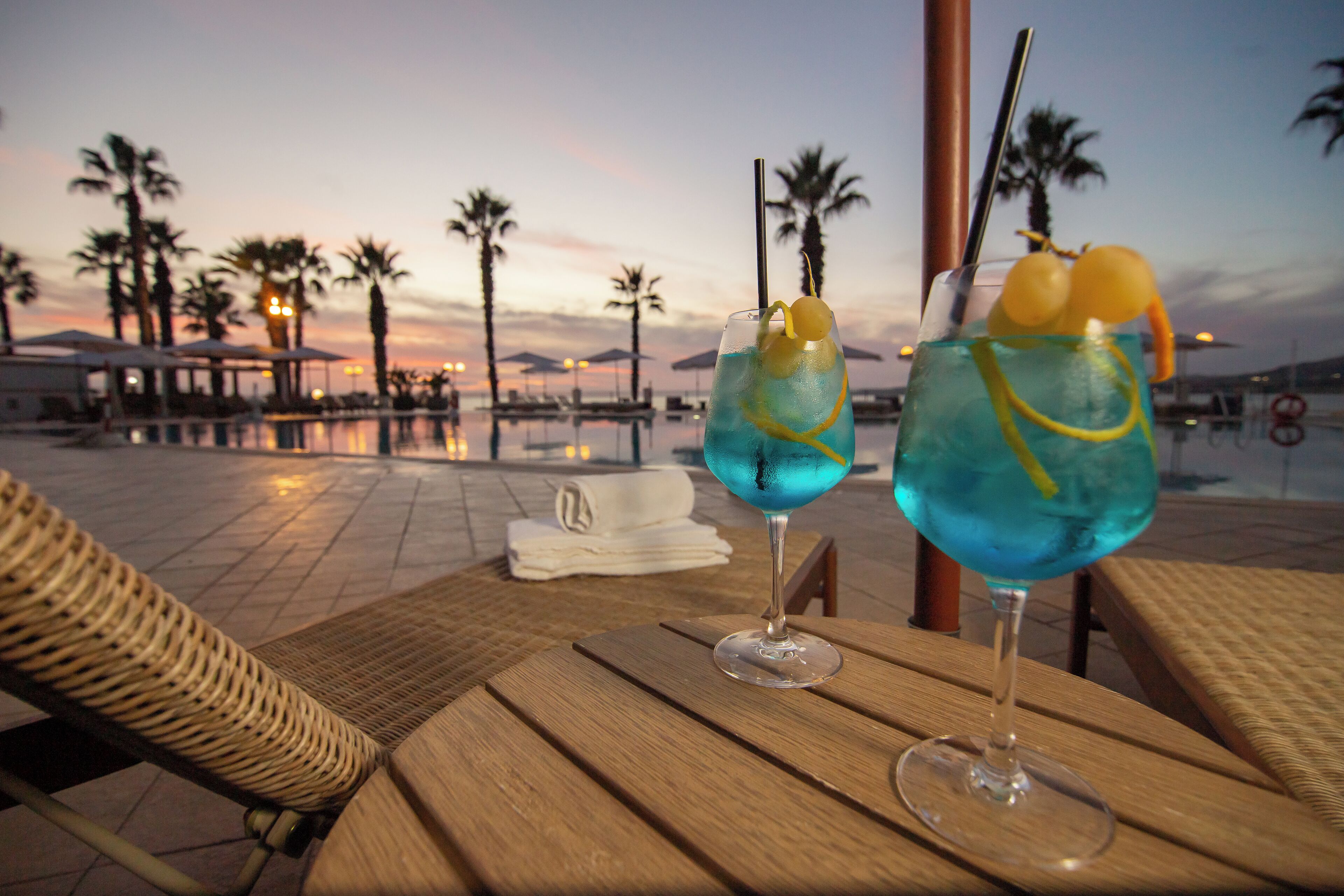 Two cocktails on a wooden table with a pool and palm trees in the background.