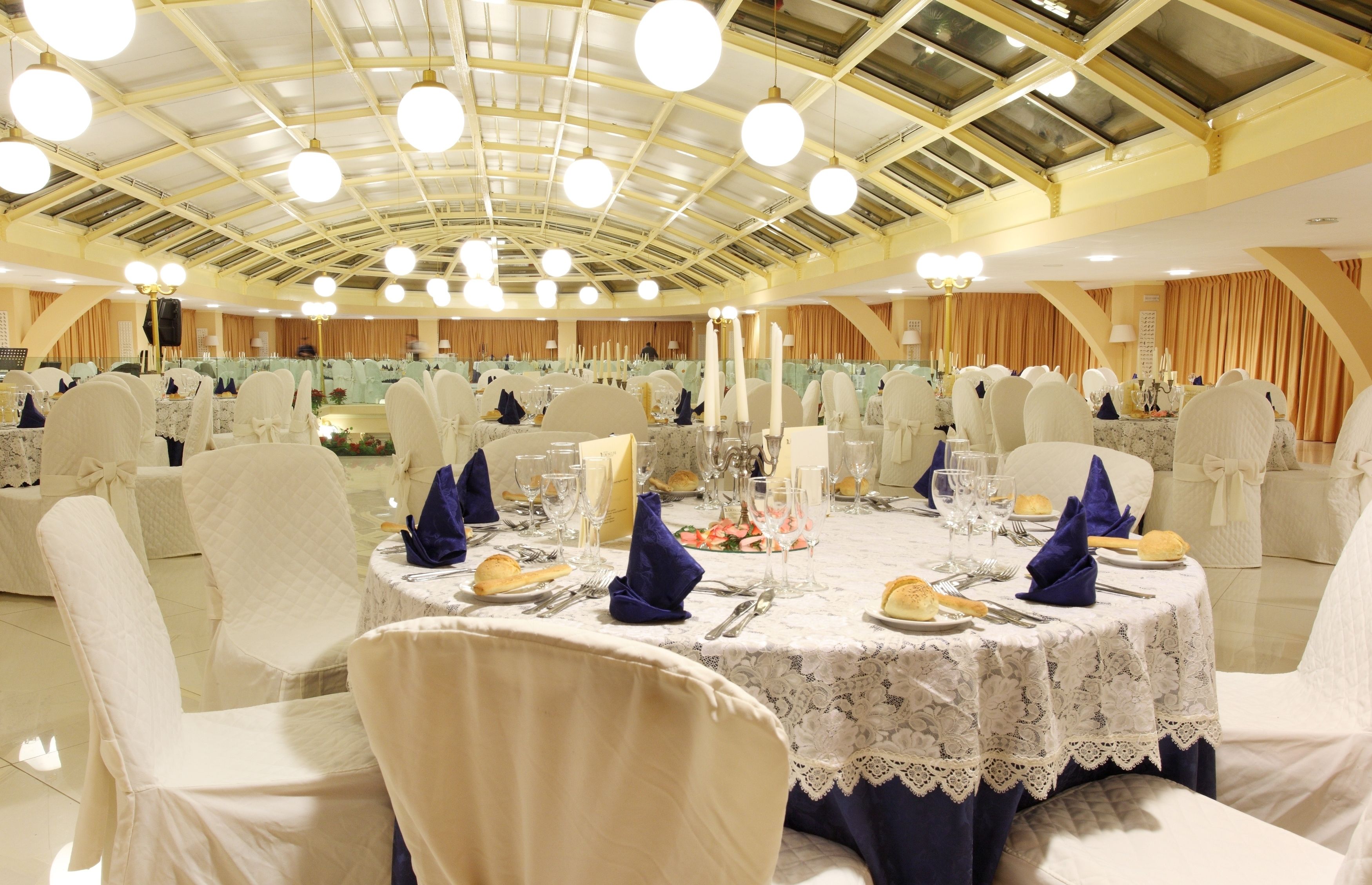 Une salle de banquet avec des tables rondes dressées pour le repas, des chaises blanches et des lustres.
