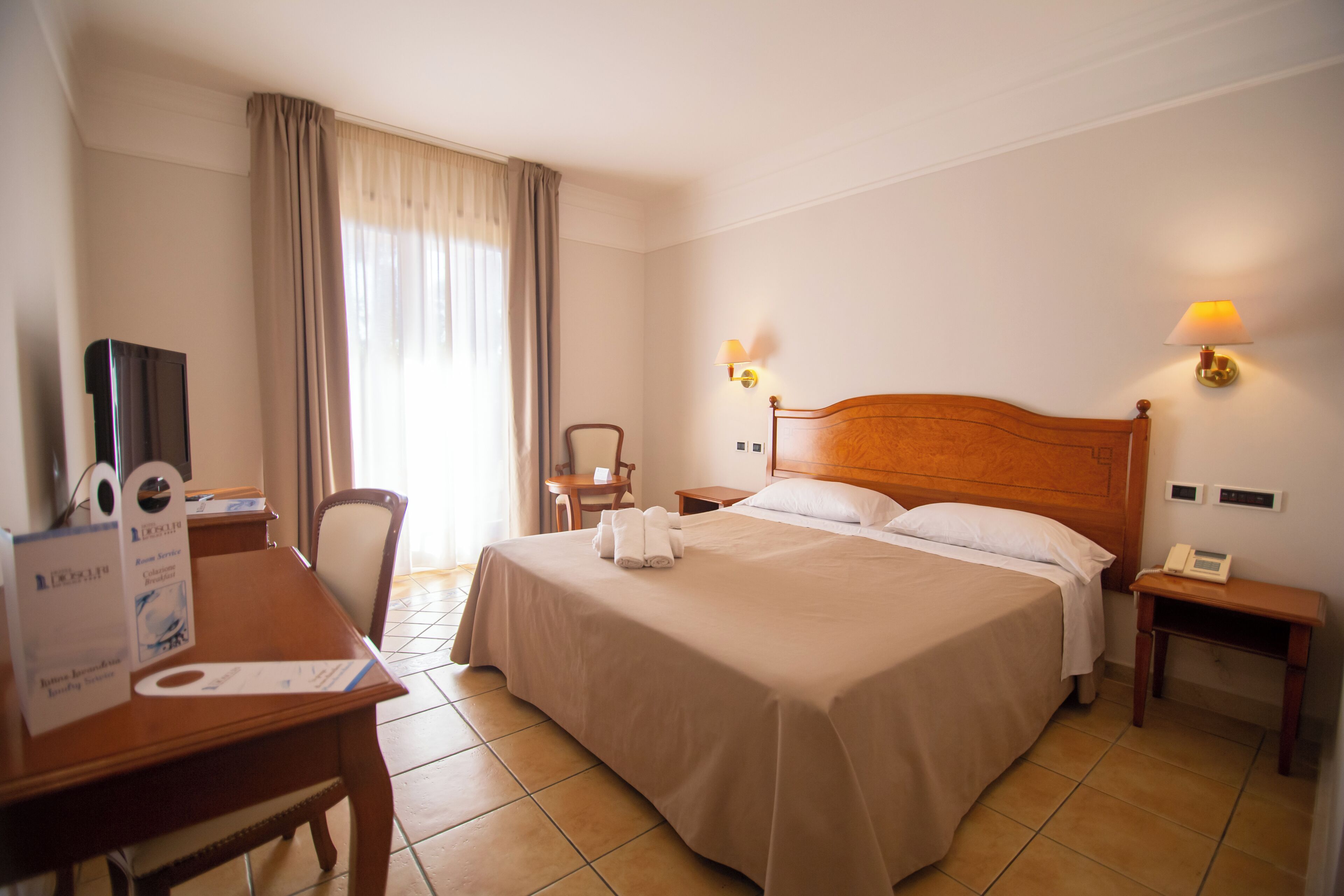 Una habitación de hotel con cama, escritorio, silla, televisión y ventana con cortinas.