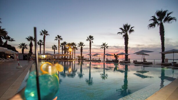 An indoor swimming pool with clear water, surrounded by palm trees and umbrellas, reflecting the sunset. - Dioscuri Bay Palace Hotel (Agrigento)