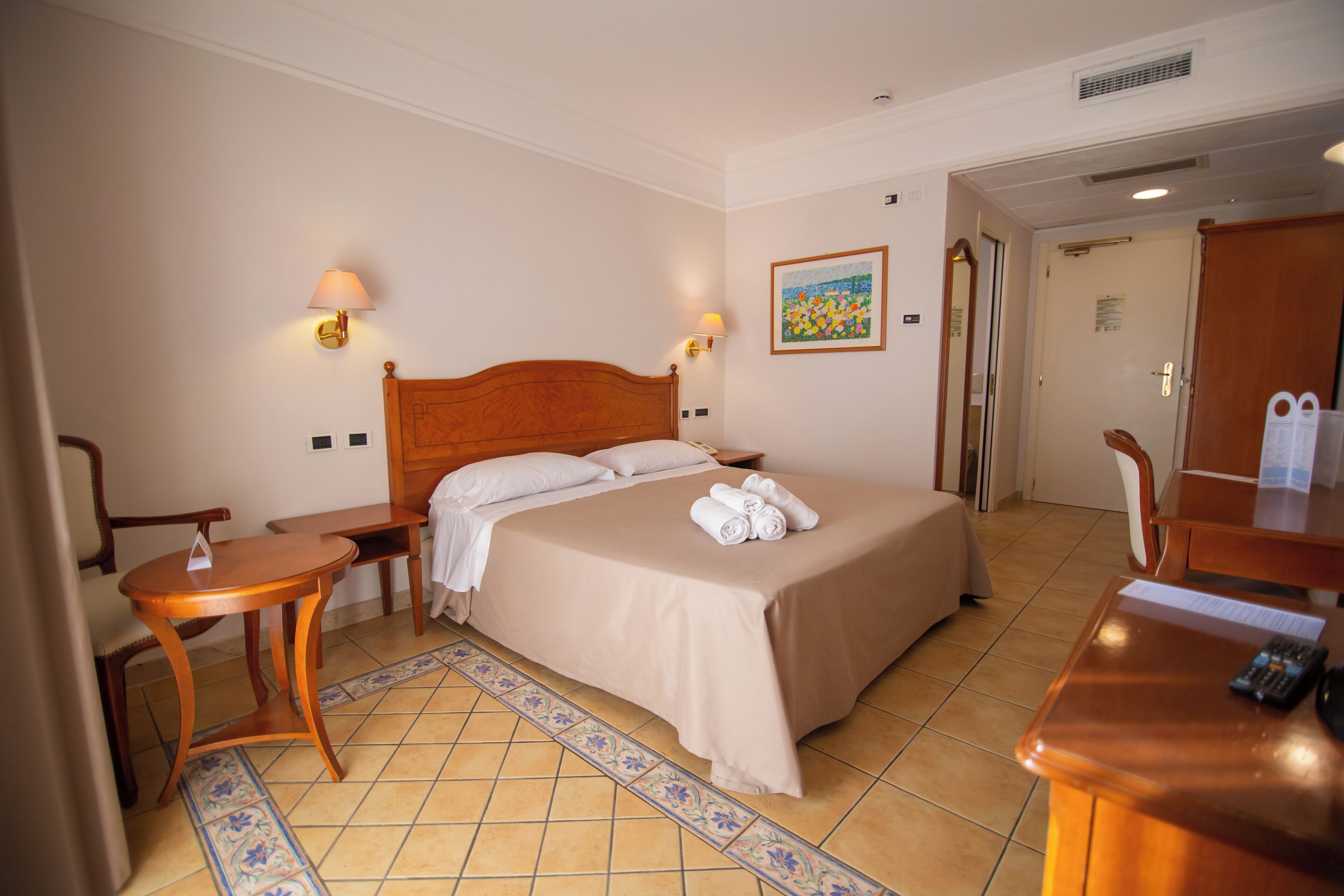Una habitación de hotel con una cama, dos mesitas de noche, un escritorio, una silla, una lámpara y un cuadro en la pared.