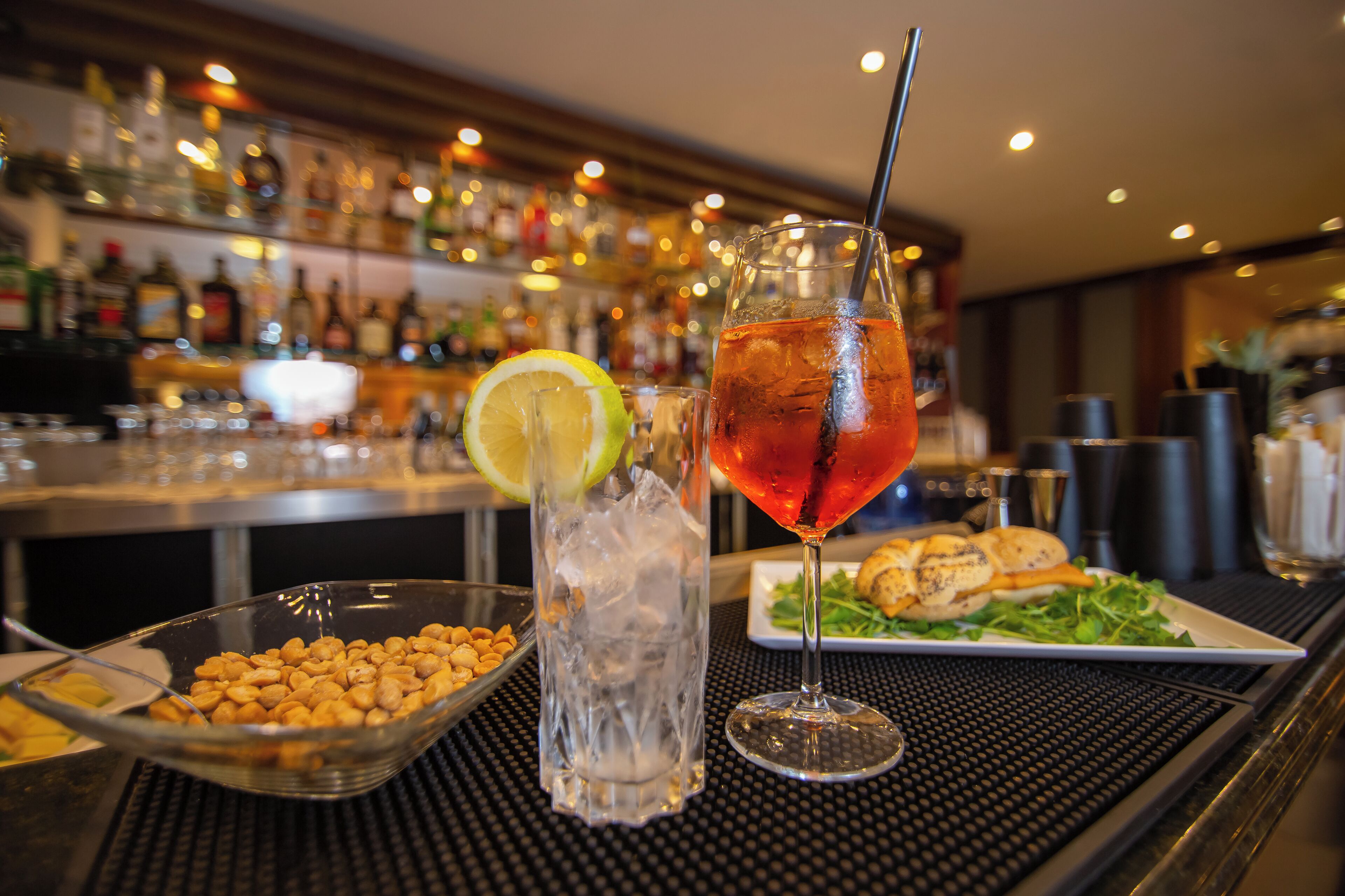 A bar setting with a glass of iced drink, a bowl of nuts, and a plate of food.