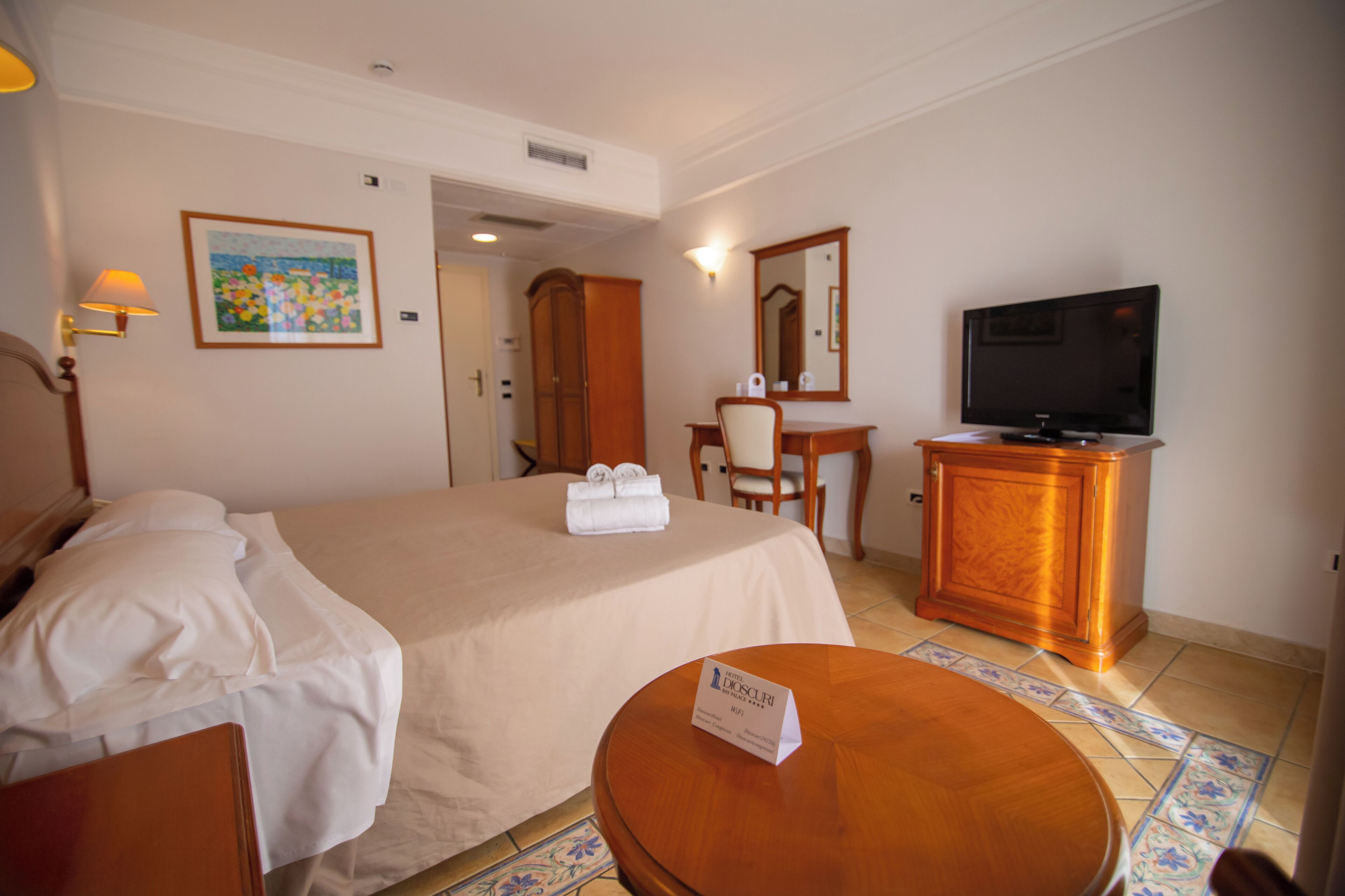 A hotel room with a bed, a desk with a chair, a television, and a small round table with a sign.