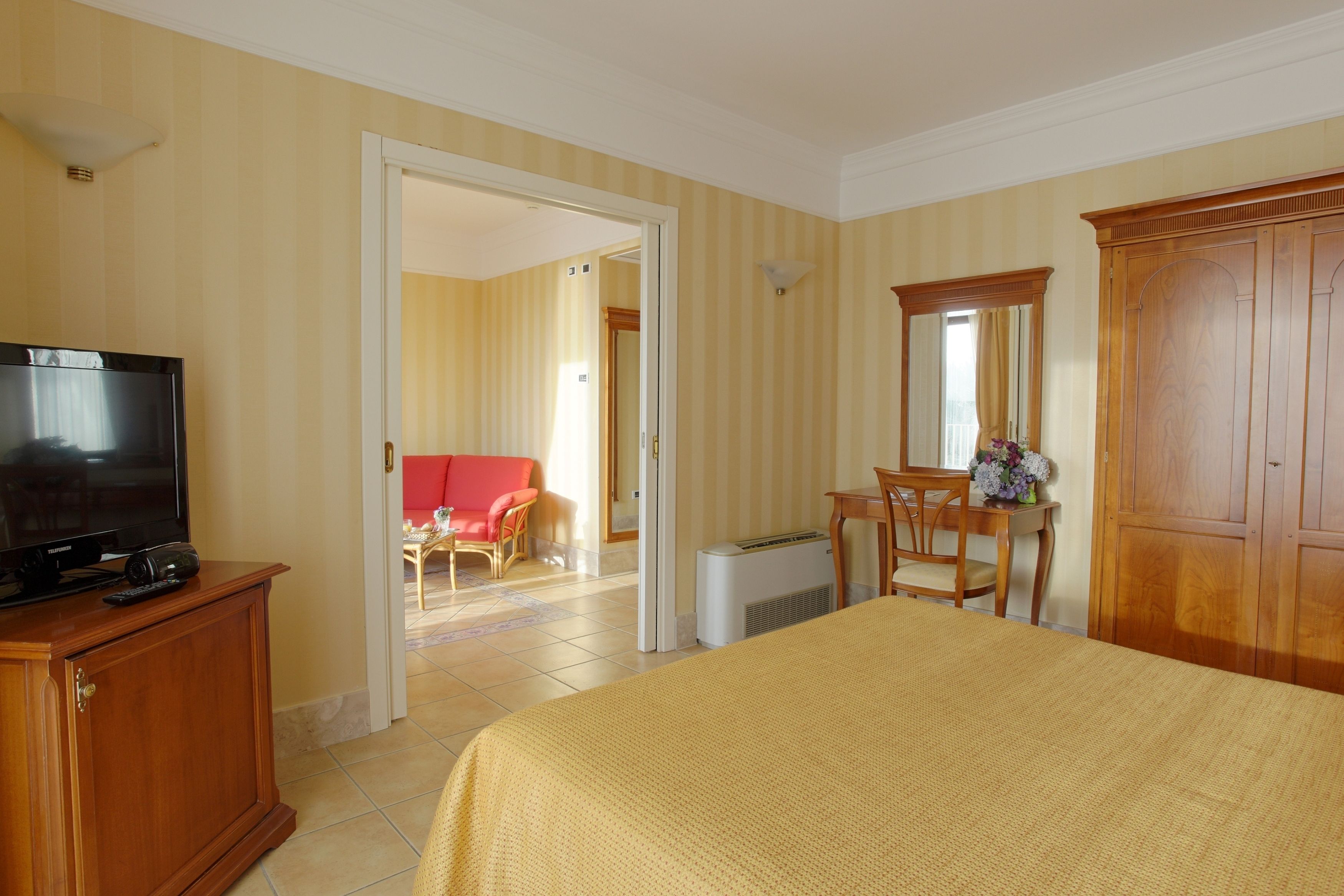 Una habitación de hotel con una cama, un televisor sobre un mueble de madera, un escritorio con una silla, un armario y una puerta que da a otra habitación.
