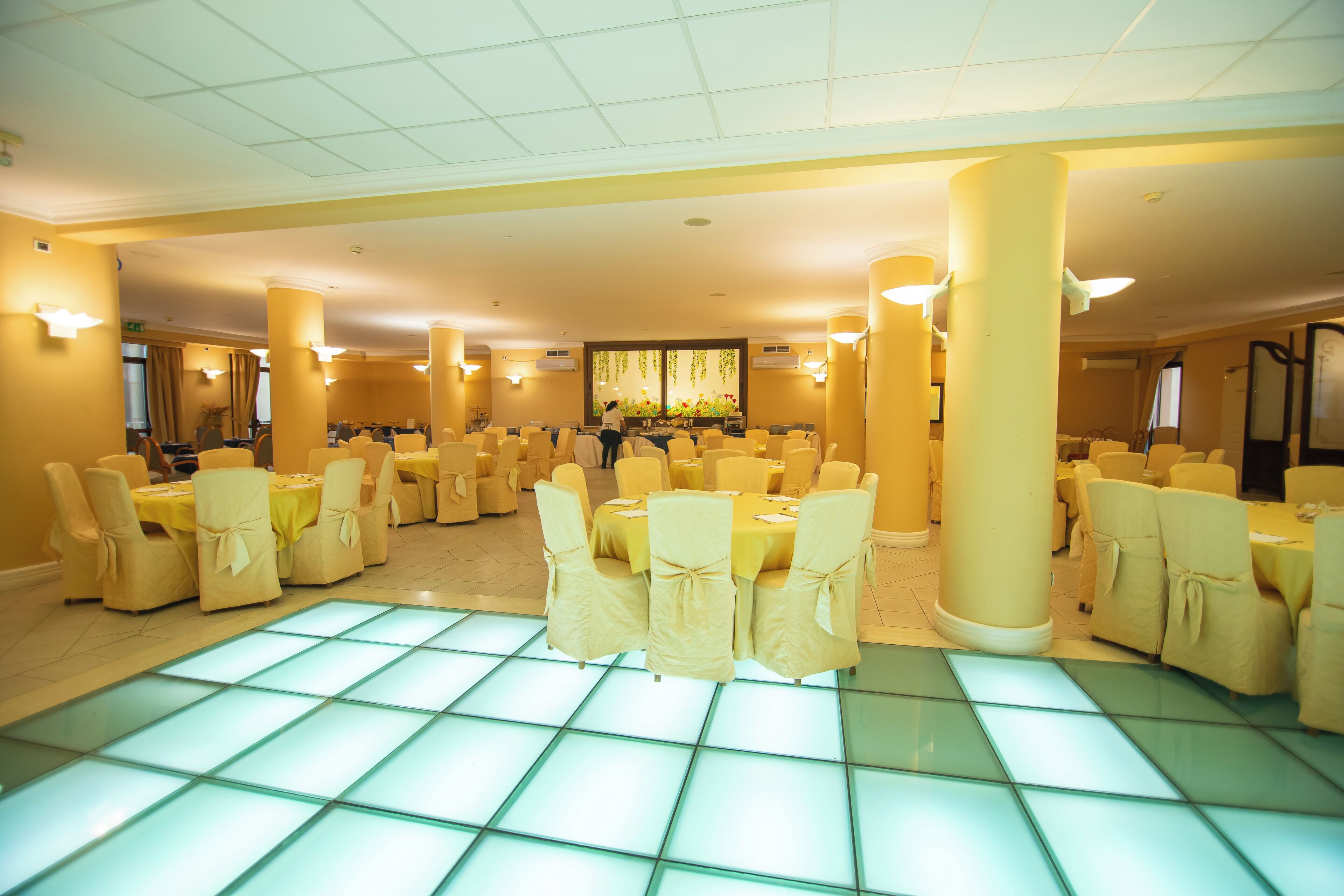 A spacious banquet hall with multiple round tables and chairs, large pillars, and a tiled floor.