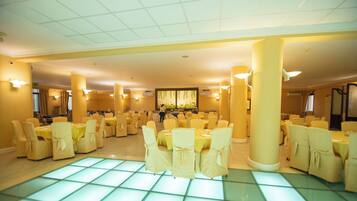 A spacious banquet hall with multiple round tables and chairs, large pillars, and a tiled floor.