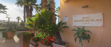 A sign for Hotel D'Isidori Bay Palace with potted plants and palm trees in the foreground.