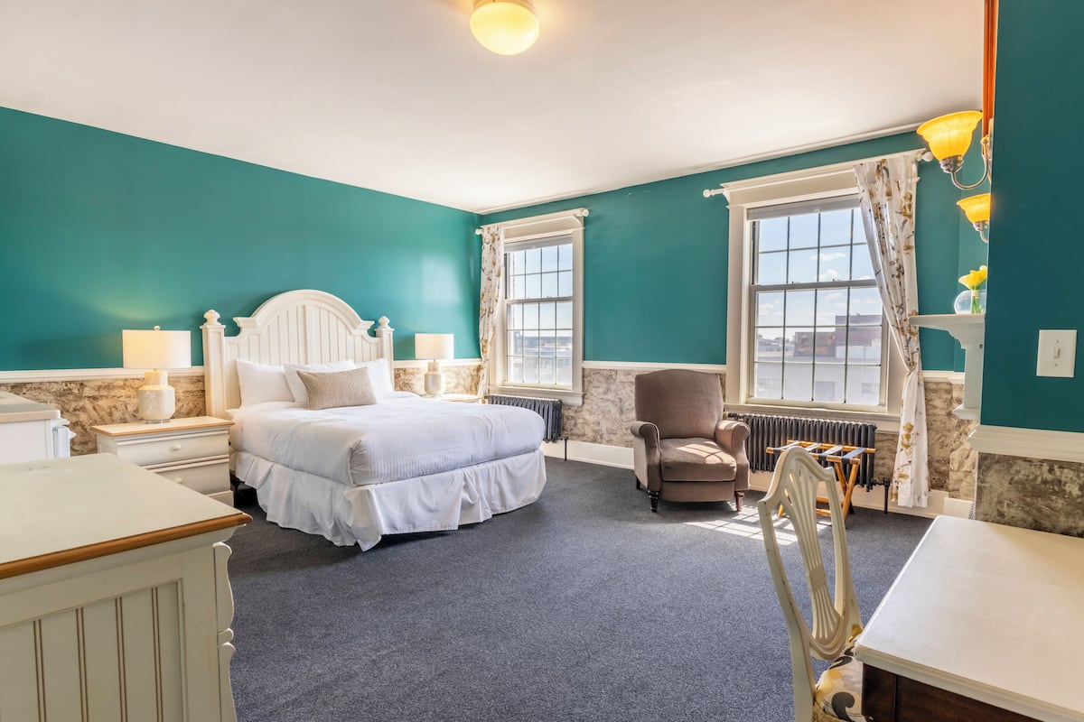 A bedroom with a large bed, a chair, a table, and two windows with curtains.