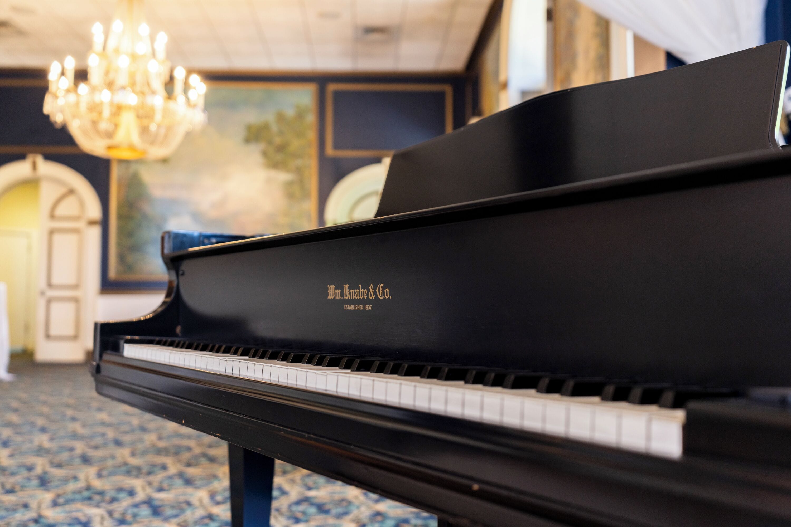 Un piano à queue au vernis noir, orné de lettres dorées, placé dans une pièce dont le sol est recouvert d’un tapis à motifs et qui est illuminée par un lustre.