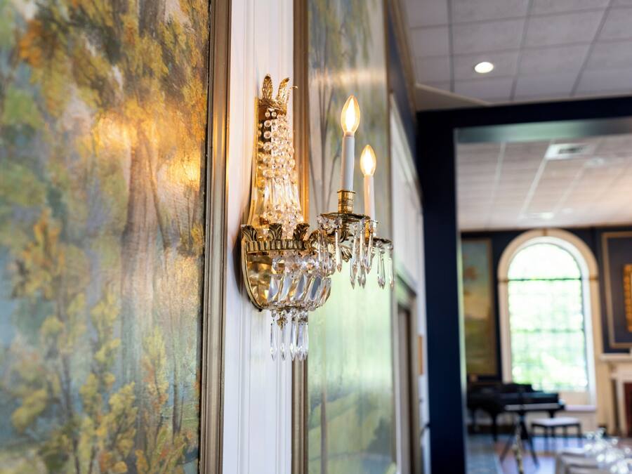 A chandelier with multiple candles, a painting on the wall, and a view of a room with tables and chairs.