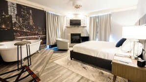 A modern hotel room with a large bed, a sitting area with a chair, a bathtub, a fireplace, and a cityscape mural on the wall. - Inn on Broadway (Rochester)