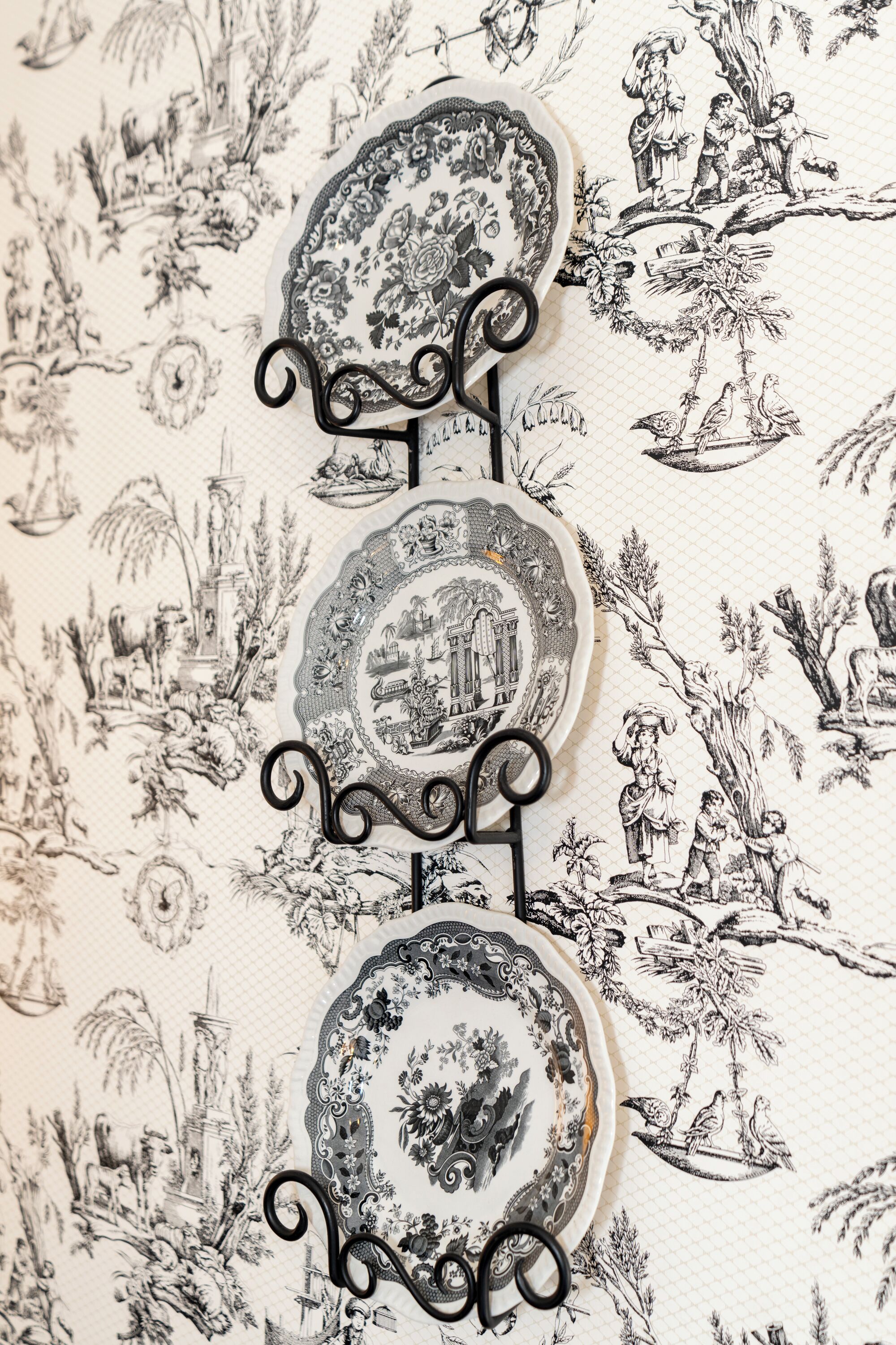 Un présentoir métallique décoratif, sur lequel sont disposées trois assiettes ornées de motifs complexes, le tout se détachant sur un fond constitué d’illustrations détaillées en noir et blanc.