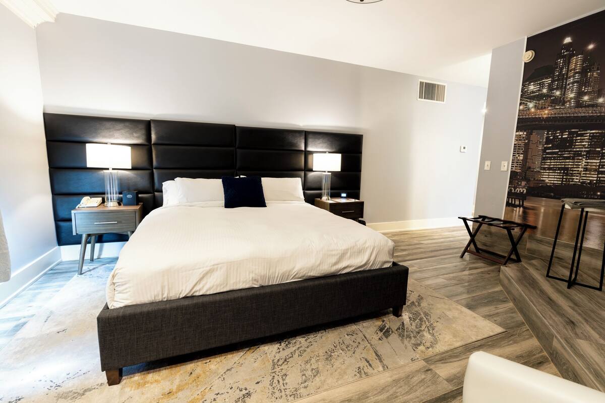 A modern hotel room with a large bed, two bedside tables, a nightstand, a chair, and a cityscape view on the wall.
