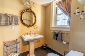 A bathroom with a pedestal sink, a gold-framed mirror, a window with curtains, and a towel rack.