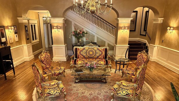 A grand living room with ornate furniture, a large chandelier, and a staircase leading to an upper floor. - Inn on Broadway (Rochester)