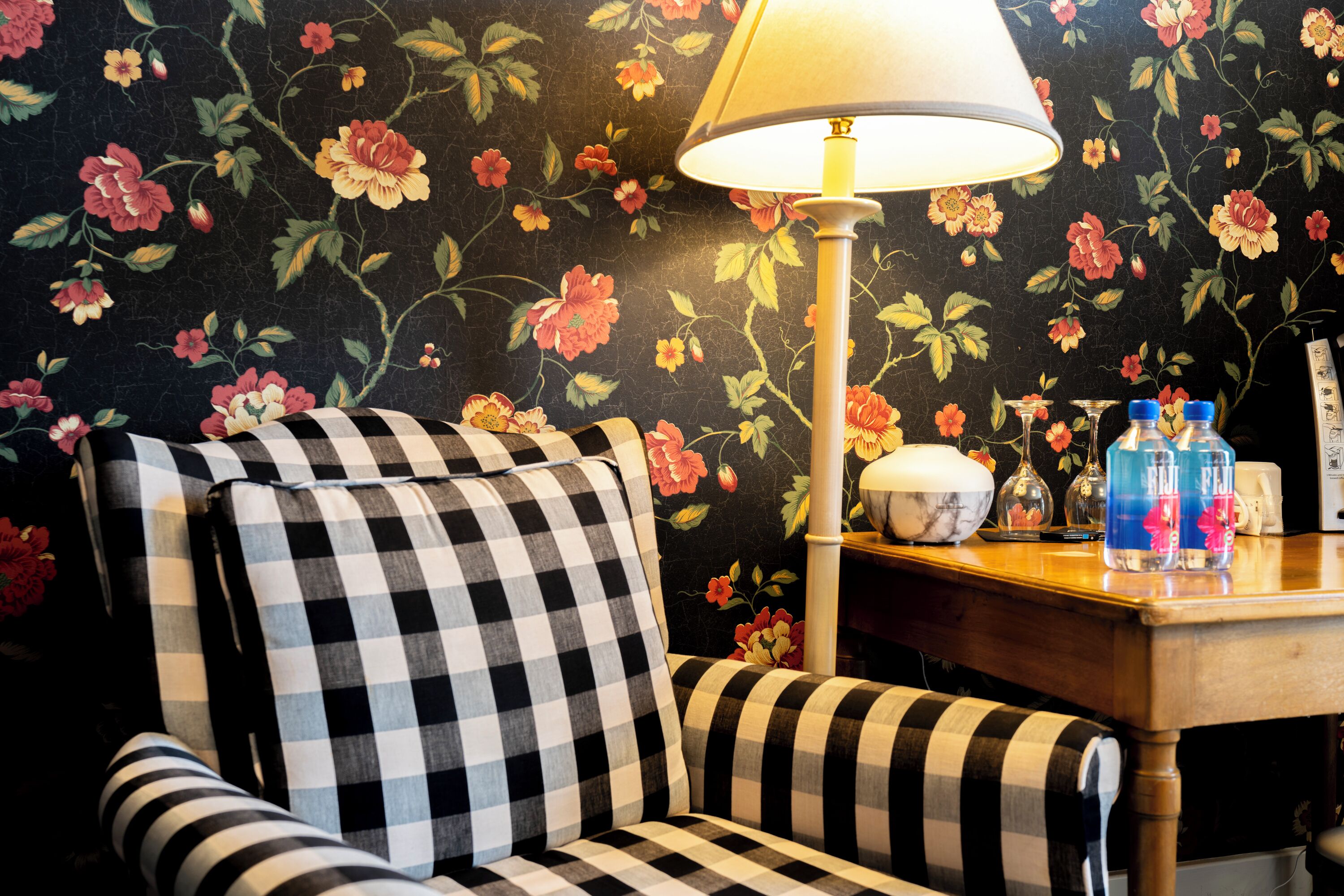 A checkered armchair with a floral wallpaper background, a table with a lamp, and decorative items.