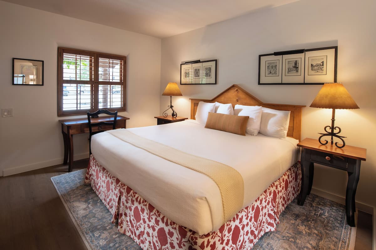 A neatly arranged bedroom with a large bed, two bedside tables with lamps, a desk, and a window with blinds.