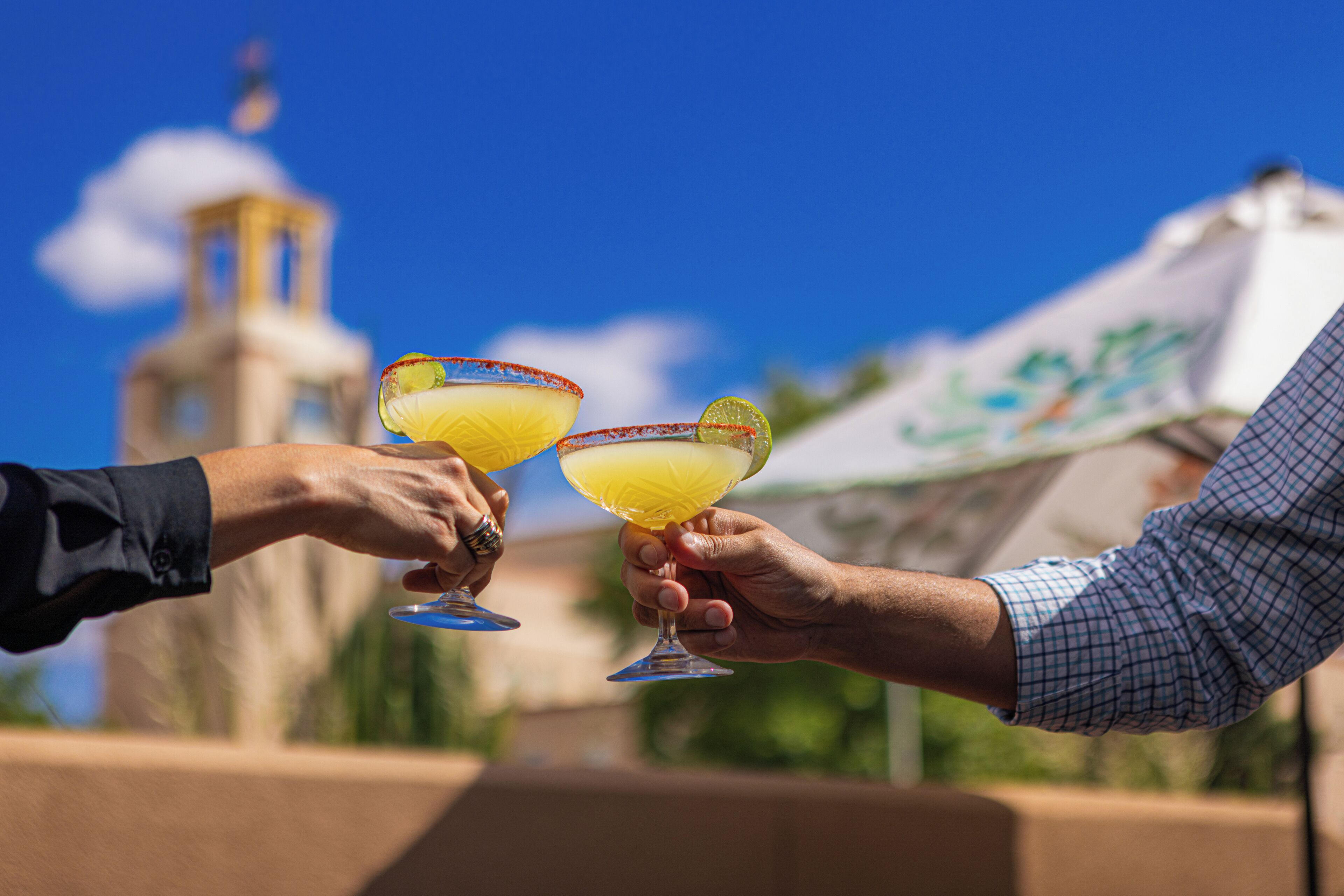 Two people holding cocktail glasses with lime garnishes, against a backdrop of a clear blue sky and a building with a dome.