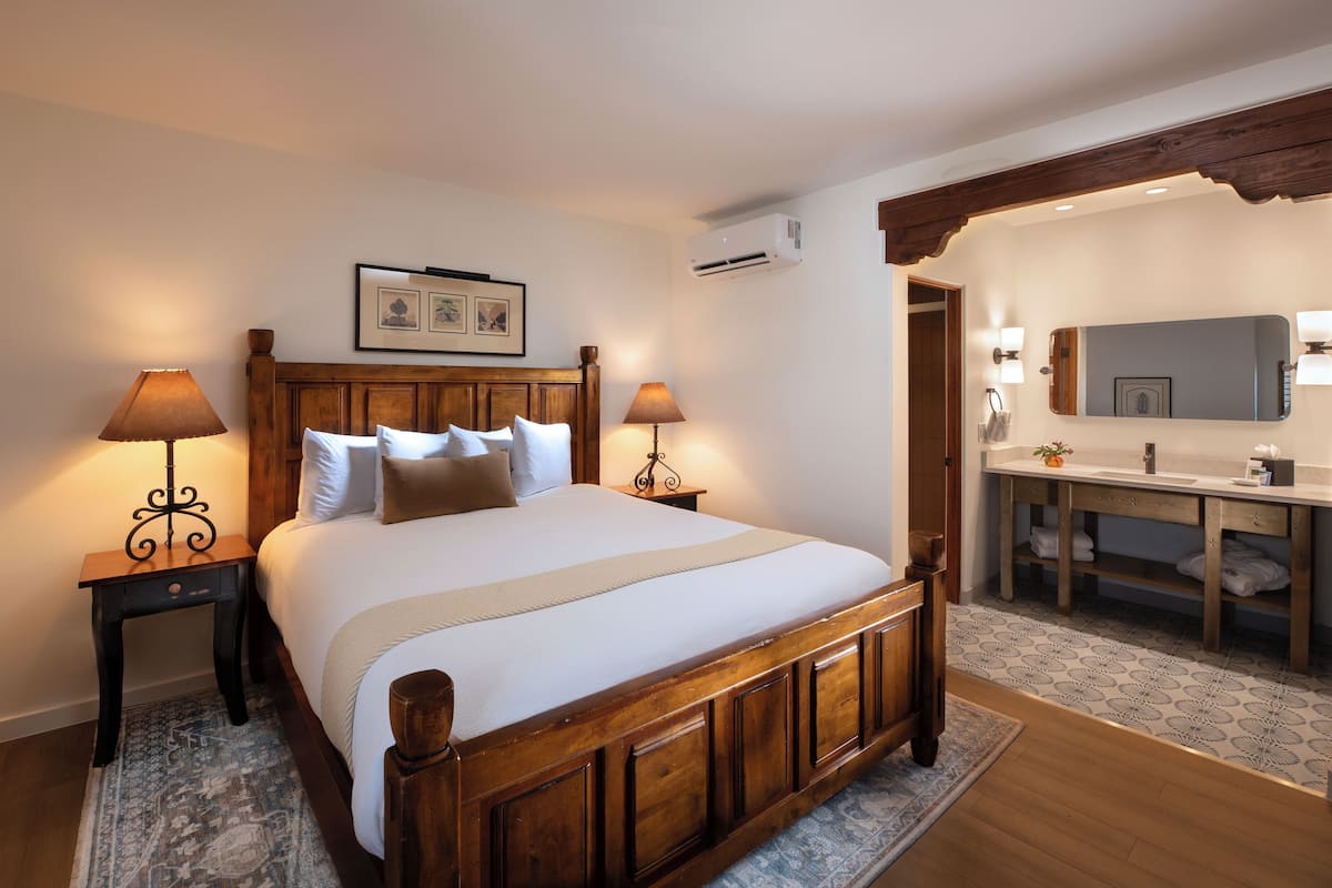A bedroom with a wooden bed, two bedside tables with lamps, a nightstand, a large mirror, and a bathroom with a sink and vanity.