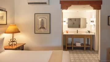 A hotel room with a bed, a bedside lamp, a framed picture, an air conditioner, a wooden ceiling, a bathroom with a sink, mirror, towel, and patterned floor.