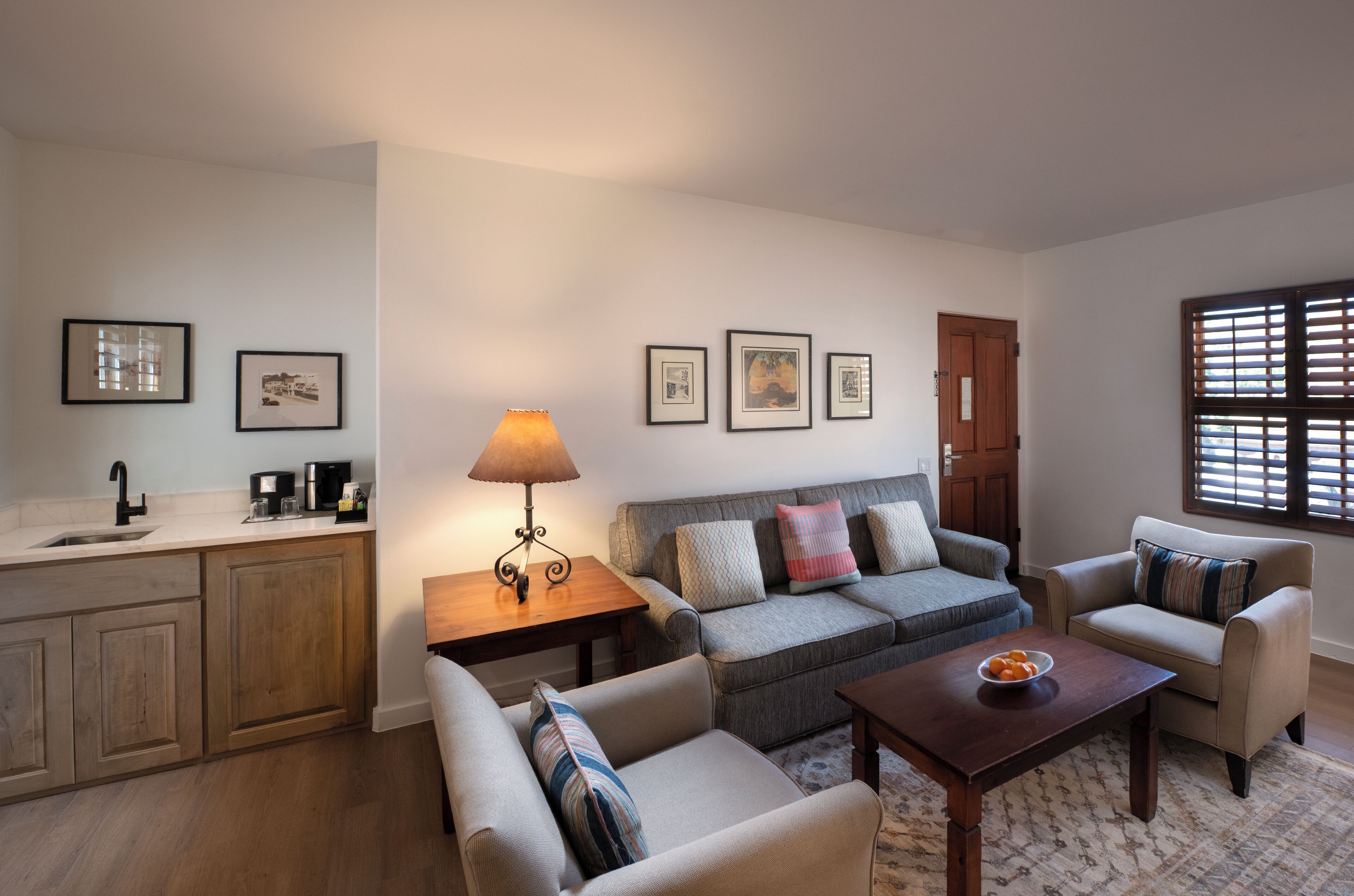 Una sala de estar con un sofá, dos sillones, una mesita de centro, una lámpara y una zona de cocina con un lavamanos y armarios.