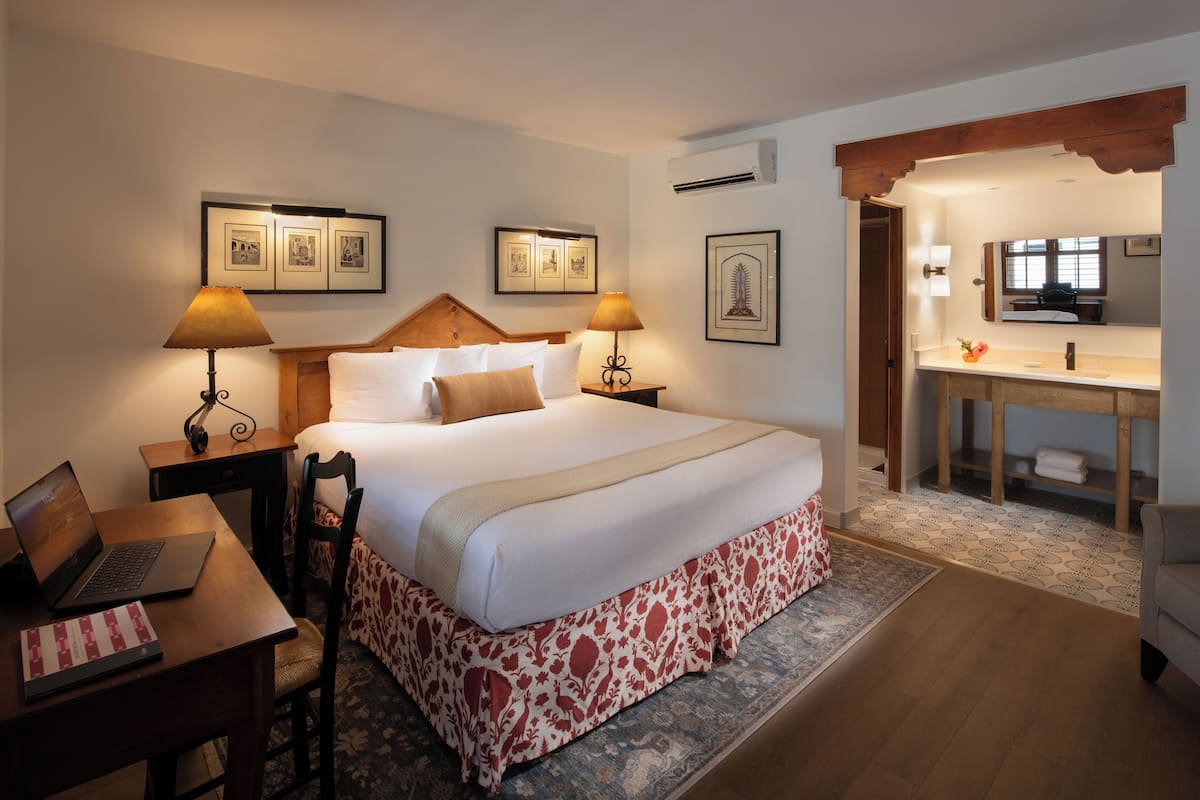 A hotel room with a large bed, two bedside tables with lamps, a desk with a laptop, and a bathroom with a mirror and sink.