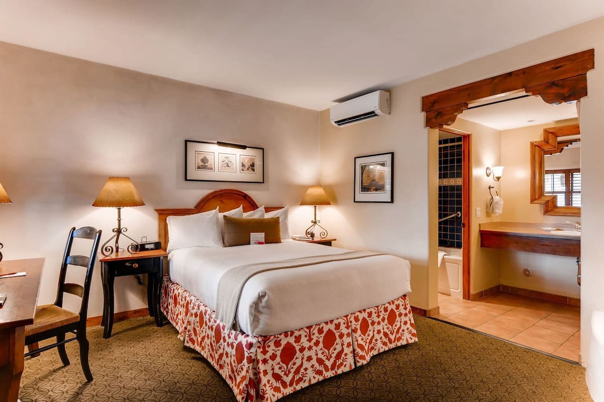A hotel room with a bed, two bedside lamps, a chair, a desk, a wall-mounted air conditioner, framed pictures, and a bathroom visible through an open door.