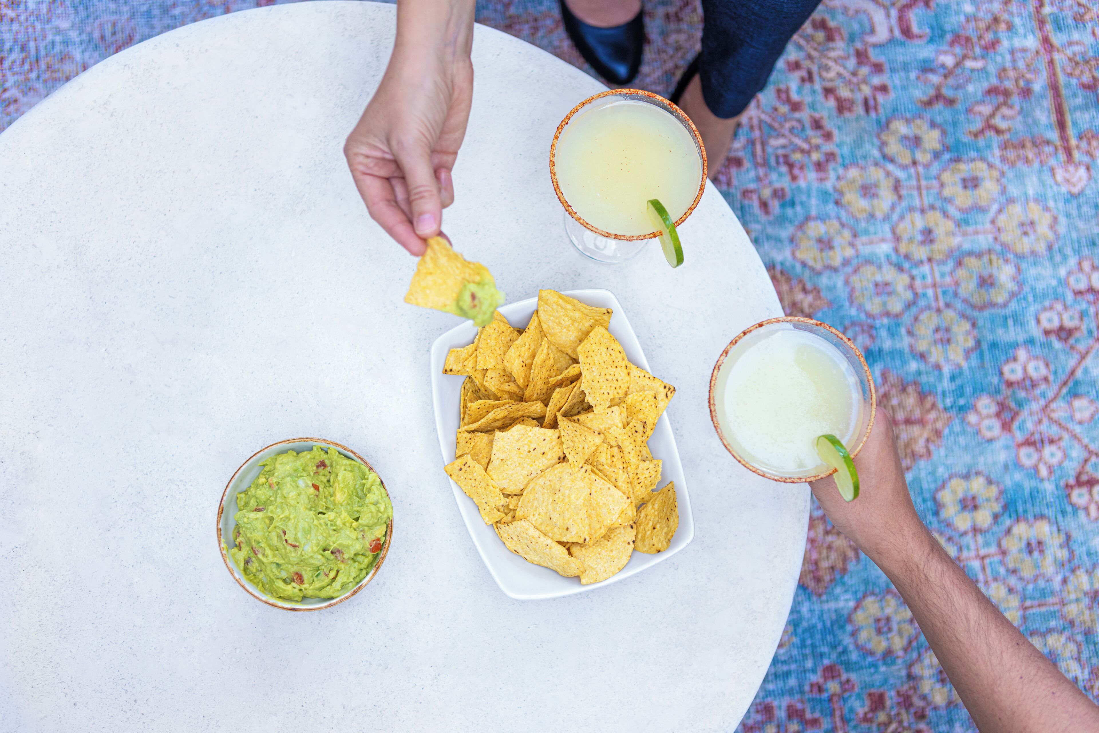 A table with a bowl of guacamole, a plate of tortilla chips, and two margarita glasses.