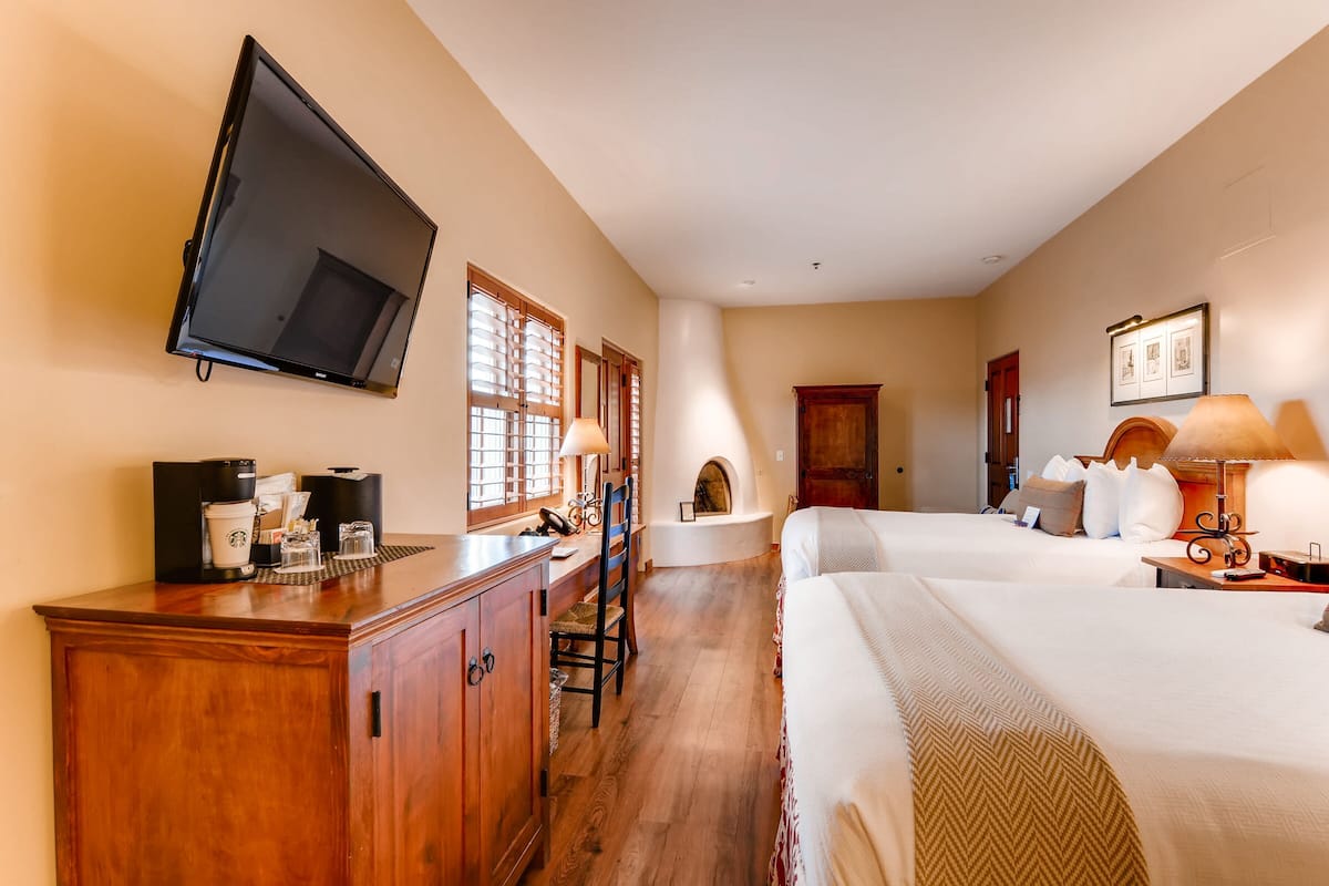 A hotel room with a flat-screen TV, a desk with a coffee maker, two beds, a fireplace, and a nightstand with a lamp.