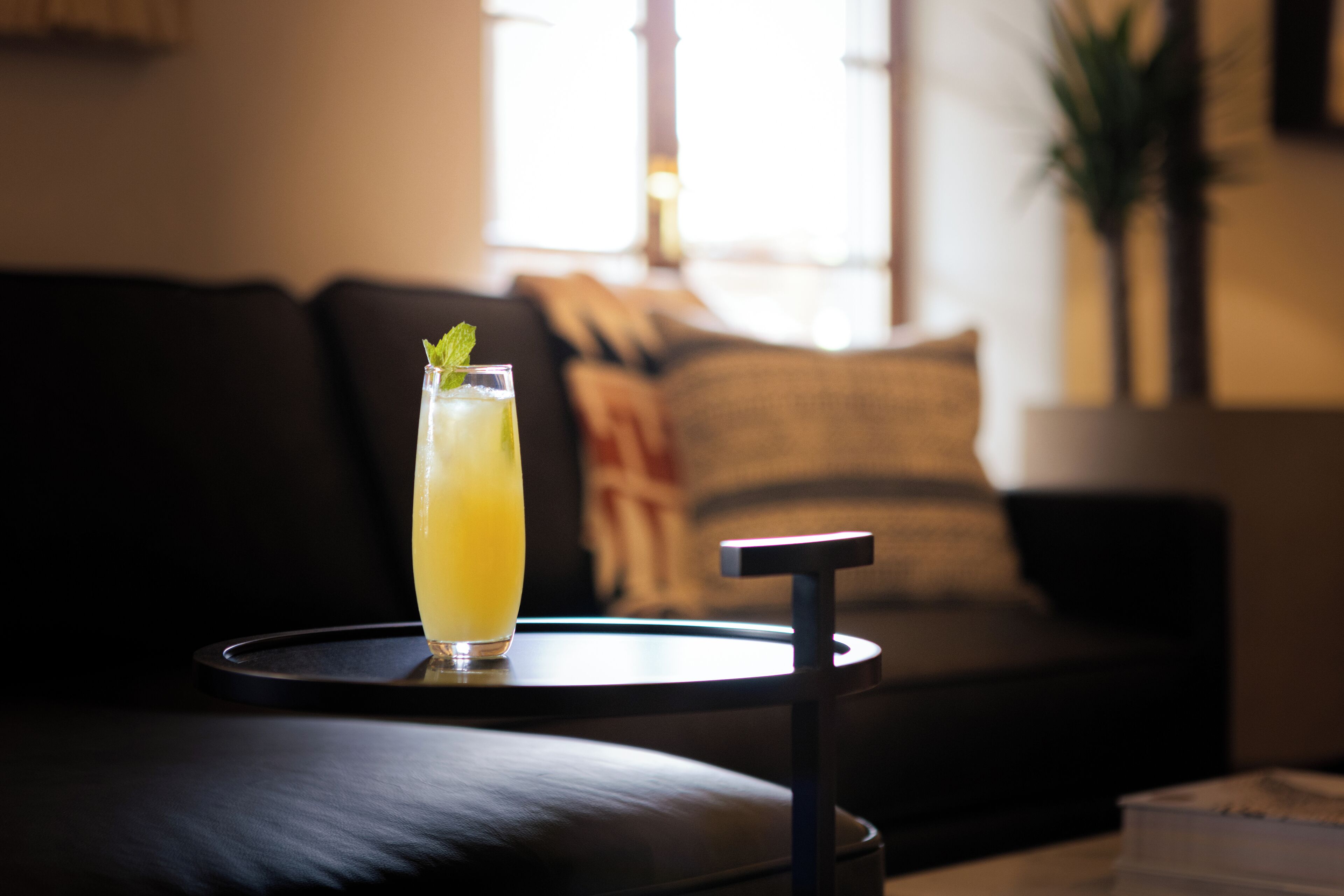 A glass of yellow beverage on a small round table in a dimly lit room with a sofa and a plant in the background.