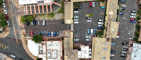 An aerial view of a parking lot with multiple rows of parked cars, a central building with a flat roof, and surrounding greenery.