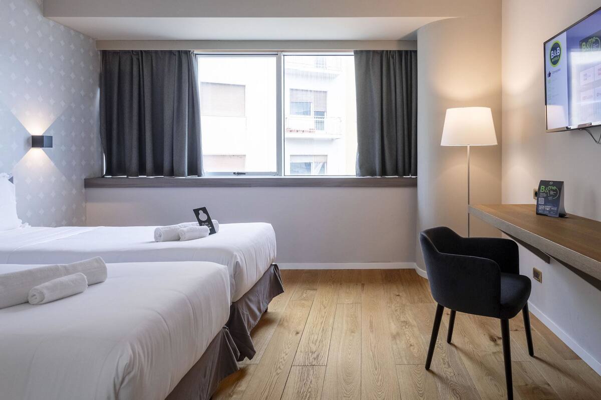 A hotel room with two beds, a desk, a chair, a lamp, and a window with curtains.