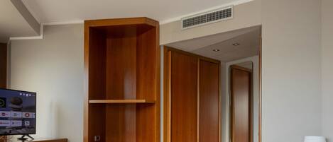 Una habitación de hotel con una cama, un televisor sobre un soporte, un armario de madera, un mueble blanco pequeño y una lámpara de noche.