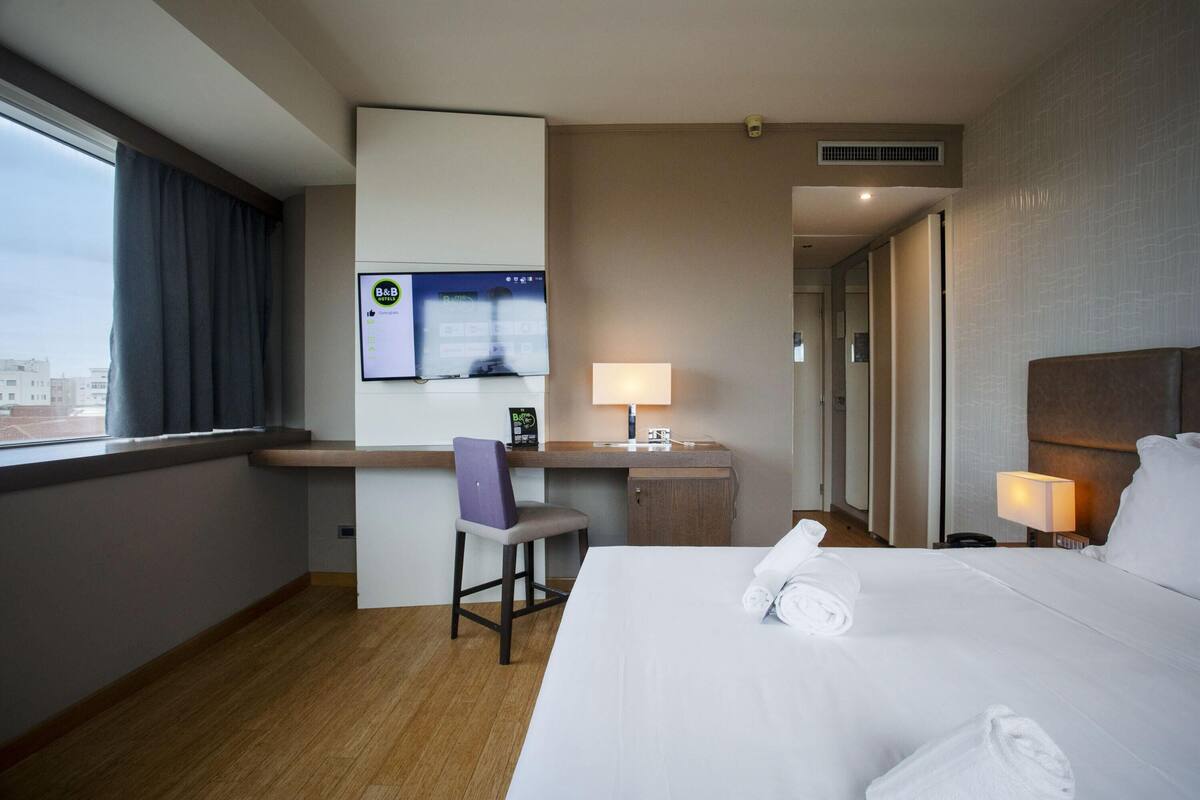 A hotel room with a bed, a desk, a chair, a TV, and a window with curtains.