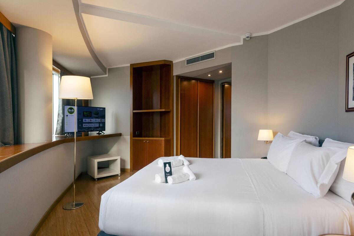A hotel room with a large bed, a desk with a computer, a wardrobe, and a door leading to another room.